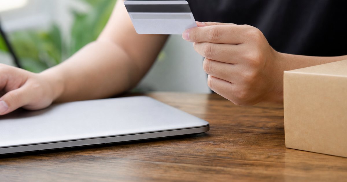 Close-up of hands holding a credit card over a wooden desk beside a closed laptop and a small cardboard box, shallow depth of field, daylight