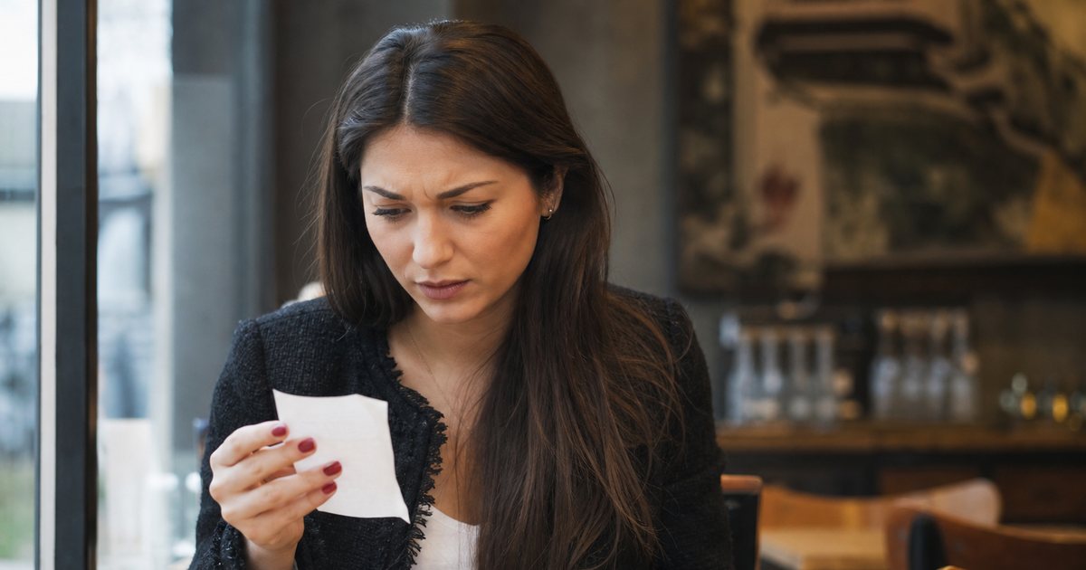 A young woman at a cafe table frowning slightly while holding a paper receipt, phone face down beside a coffee cup, daylight through a window