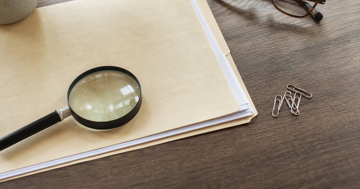 Overhead view of a tidy desk with a manila folder, paper clips, a magnifying glass, and a pair of reading glasses, natural light